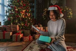 Merry Christmas. African American woman unpacking open up gift box near Christmas tree. Girl in living room with Christmas tree and fireplace opening gift box with surprise face, Christmas eve at home photo