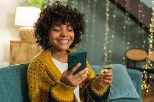 mujer afroamericana comprando en línea con un teléfono inteligente pagando con tarjeta de crédito dorada. niña sentada en casa comprando en internet ingrese los detalles de la tarjeta de crédito. servicio de entrega de comercio electrónico de compras en línea. foto