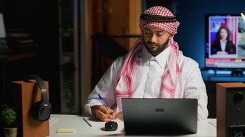 retrato de sonriente árabe empresario trabajando a hogar oficina escritorio en computadora portátil, hojeada el Internet y escritura en cuaderno. medio oriental hombre utilizando bolígrafo, bloc y digital dispositivo mientras teletrabajo video