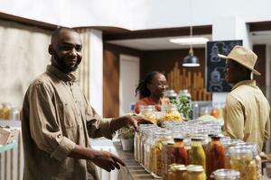 A smiling African American man looking at the camera in a modern grocery store. Reusable packaging, eco friendly products, and a variety of items are showcased on the shelves. photo