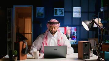 Muslim teleworker remotely working in dimly lit apartment, enjoying fresh cup of coffee. Middle Eastern worker at home typing on notebook device, sending emails and drinking hot beverage video