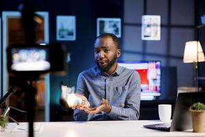 African American tech enthusiast presents latest smart light bulb to audience while discussing home improvement in video on online channel, highlighting wonders of WiFi home automation photo