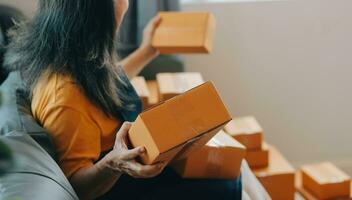 Startup SME small business entrepreneur of freelance Asian woman wearing apron using laptop and box to receive and review orders online to prepare to pack sell to customers, online sme business ideas. photo