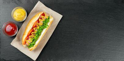Homemade Hot Dog with mustard, ketchup, tomato and fresh salad leaves on black slate background photo