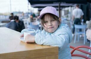 divertido linda niña en calle cafetería. pequeño niña Bebiendo caliente bebida foto