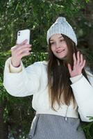 adolescente niña utilizando Teléfono móvil en bosque, haciendo selfie dispara vídeo retrato, ondulación mano, diciendo Hola foto
