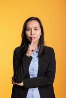 Portrait of filipino woman making shh gesture in studio by putting forefinger over lips posing over yellow background. Silent adult shushing people gesturing secrecy and confidentiality. photo