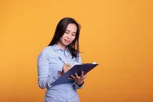 Serious asian woman writing business report on documents, analyzing questionnaire and answering question in studio. Confident model standing over yellow background looking at checklist photo