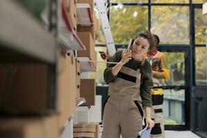 Storage room worker having remote conversation with supervisor, talking at landline phone in storage room. Woman working at products inventory, checking boxes with clients orders preparing shipment photo