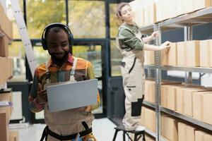 Storage room employee listening music at headset, checking clients shipping details on laptop computer. African american supervisor working at products distribution, preparing goods in storehouse photo