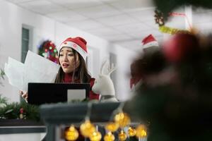 Businesswoman overwhelmed by stressful company project during Christmas holiday season. Frustrated workaholic employee struggling to finish overload paperwork in festive decorated office photo