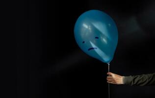 mental salud trastorno concepto. un estresado, ansiedad, Deprimido persona participación un azul globo con un tristeza cara desde el oscuro , negativo emoción y sentimiento. malhumorado. oscuro tono foto