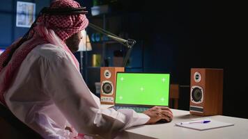 Muslim man scrolling on green screen laptop, checking social media feed. Self employed person surfing the internet from chroma key device in apartment office with TV as background noise video