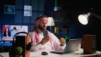 Muslim worker answering phone call while teleworking from apartment office desk. Middle Eastern company executive talking with colleague, discussing paperwork and tasks needing to be solved video