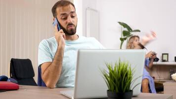 Man talks on the phone with a laptop in front while her wife is playing video games on a console in the background and eats popcorn. Dolly slider rack focus 4K footage