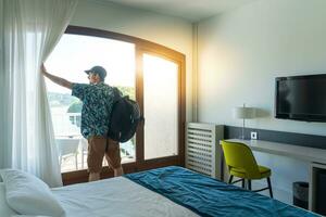 turista hombre en el hotel dormitorio con su equipaje en pie cerca el ventana. viaje y vacaciones concepto. foto