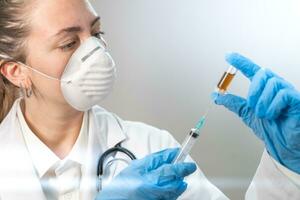 Young attractive female doctor preparing vaccine syringe wearing preventive mask and blue gloves. Coronavirus. photo