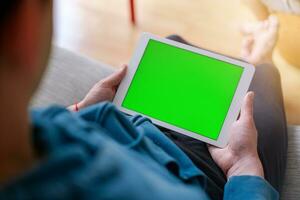 Over the shoulder view of a man working on a tablet with a green chroma key screen while sitting on a couch photo