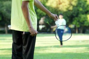 An Asian boy holds a badminton racket and a white shuttlecock while playing badminton with friends on the park lawn in the evening after returning from school. photo