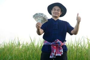 Handsome Asian man farmer is at paddy field, holds cash Thai banknote money, thumbs up. Feels confident. Concept, Agriculture occupation. Thai farmer. Profit, income. photo