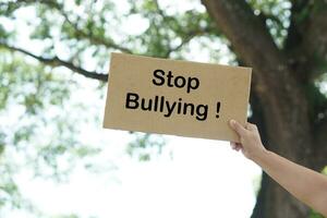 Close up hand holds paper placard with text Stop Bullying, outdoor background.  Concept, campaign for calling everybody to stop bullying each other by using bad word, action, texting or online. photo