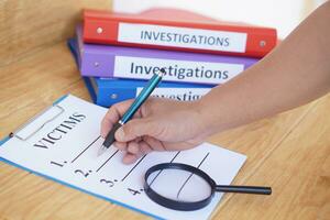 Close up detective hand is writing victims list on paper notepad clipboard. magnifying glass and investigation binders. Concept, crime. Analyzing criminal investigation. photo