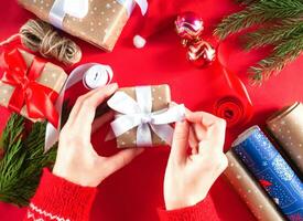 Woman's hands packing a gift box. Christmas background with paper, gift boxes, ribbon, fir branches, twine, decor on the red background. Preparation for holidays. Christmas or New Year flatlay. photo