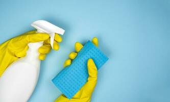 Woman's hands in rubber gloves hold a cleaner and rag on a blue background. Housekeeping concept. Close-up. Copy space. Top view. photo