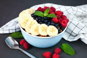 Oatmeal bowl with fresh raspberries, blueberries and ripe banana on a dark background. Healthy and tasty breakfast. photo