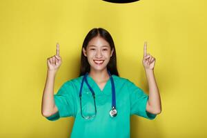 retrato de un hermosa joven mujer en un amarillo fondo, simpático hermosa mujer vistiendo un del doctor uniforme y señalando mientras vistiendo un estetoscopio. foto