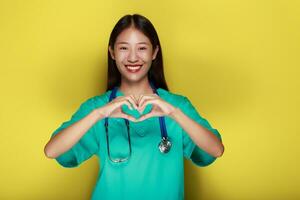 un asiático mujer vistiendo un del doctor uniforme poses con su manos en el forma de un corazón en pie en frente de un amarillo antecedentes. foto