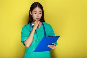 Beautiful young woman makes a thoughtful expression while wearing a doctor's uniform standing in front of a yellow background. photo