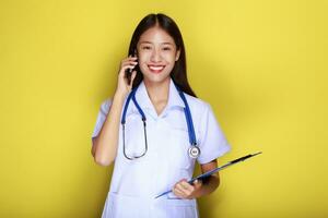 retrato de un hermosa joven mujer en un amarillo fondo, asiático mujer poses con un célula teléfono mientras vistiendo un del doctor uniforme y un estetoscopio. foto