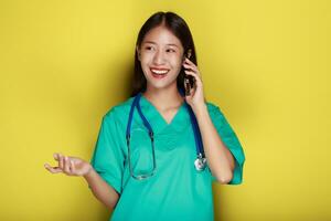 retrato de un hermosa joven mujer en un amarillo fondo, asiático mujer poses con un célula teléfono mientras vistiendo un del doctor uniforme y un estetoscopio. foto