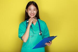 Beautiful young woman makes a thoughtful expression while wearing a doctor's uniform standing in front of a yellow background. photo