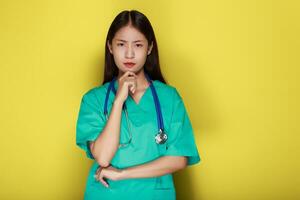 hermosa joven mujer hace un pensativo expresión mientras vistiendo un del doctor uniforme en pie en frente de un amarillo antecedentes. foto