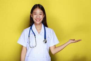 retrato de un hermosa joven mujer en un amarillo fondo, simpático hermosa mujer vistiendo un del doctor uniforme y señalando mientras vistiendo un estetoscopio. foto