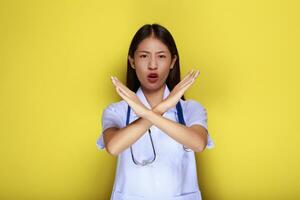 un asiático mujer vistiendo un del doctor uniforme poses con un mano en negación en frente de un amarillo antecedentes. foto