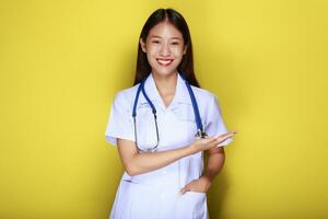 Portrait of a beautiful young woman in a yellow background,  Friendly beautiful woman wearing a doctor's uniform and pointing while wearing a stethoscope. photo