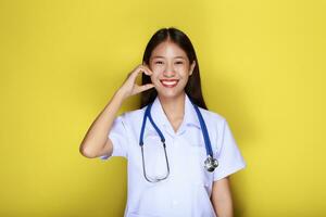 An Asian woman wearing a doctor's uniform poses with her hands in the shape of a heart standing in front of a yellow background. photo