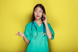 retrato de un hermosa joven mujer en un amarillo fondo, asiático mujer poses con un célula teléfono mientras vistiendo un del doctor uniforme y un estetoscopio. foto