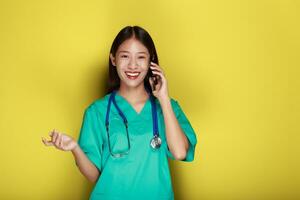 retrato de un hermosa joven mujer en un amarillo fondo, asiático mujer poses con un célula teléfono mientras vistiendo un del doctor uniforme y un estetoscopio. foto