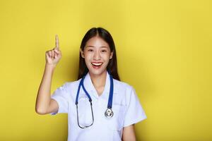 retrato de un hermosa joven mujer en un amarillo fondo, simpático hermosa mujer vistiendo un del doctor uniforme y señalando mientras vistiendo un estetoscopio. foto