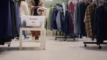 Customers donate wares in charity thrift shop where profits from vintage clothes on racks bought by clients are given to charitable causes. Generous man placing garments in clothing store donation box video