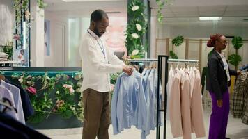 sonriente Al por menor asistente arreglando ropa en centro comercial ropa Tienda con rápido Moda atuendo siguiendo el último tendencias alegre trabajador organizando surtido de elegante hombres chaquetas americanas en Moda boutique video