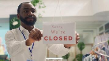 BIPOC man closing down store in order to do restocking in elegant high end fashion boutique. African american employee adding closed sign on clothing store door, adding new collection photo