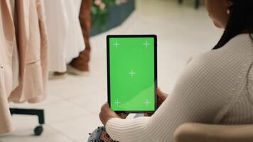 Shopper in second hand clothing shop holding green screen tablet, trying to decide what clothes to purchase. Client looking at new collection of garments in discount store using chroma key device photo