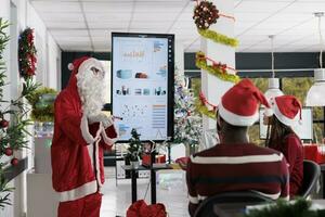 Certified lecturer dressed as Santa Claus teaching employees important business growth hacks in festive ornate workspace. Public speaker showing statistics charts to workers during holiday season photo