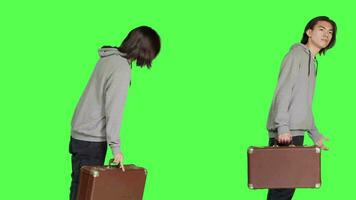 Young adult holding vintage suitcase over greenscreen backdrop, waiting impatiently for a certain thing. Asian man carrying huge briefcase while he looks at time on wrist watch in studio. video