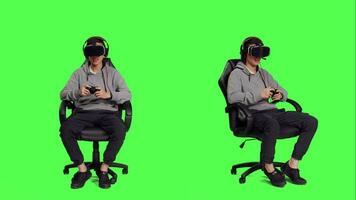 Young guy playing competition with vr headset sitting over greenscreen backdrop, enjoying video games on joystick. Asian gamer having fun with online rpg contest, virtual reality leisure.